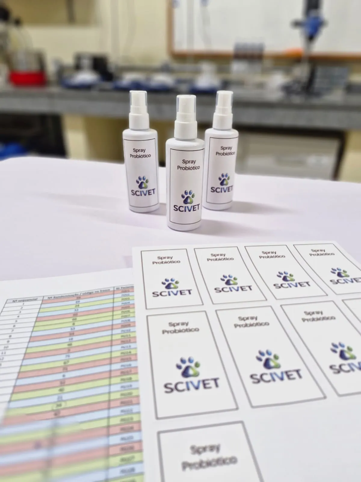 Três frascos do spray probiótico da Scivet sobre a bancada do laboratório, ao lado de folhas de rótulo impressas e de uma planilha de acompanhamento de estabilidade.